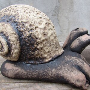 May include: A ceramic snail sculpture with a brown and beige shell and a textured surface. The snail is lying on its side with its foot extended.