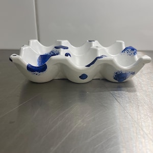 May include: A white egg carton with blue paint splatters. The carton has six individual egg holders.
