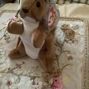 May include: A brown and white stuffed squirrel with a red and white Ty tag. The squirrel is sitting on a white fabric with embroidered flowers.