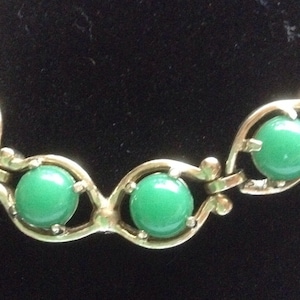 May include: A gold chain necklace with green glass stones. The chain is made of a series of linked loops with a decorative design.