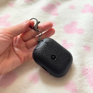 以下が含まれることがあります： 黒い革製AirPodケース、金属製の留め金付き。ケースはテクスチャ加工された表面で、前面に小さなボタンが付いています。留め金は金属リングに取り付けられています。