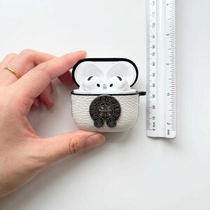 以下が含まれることがあります： 黒い縁と銀色のエンブレムが付いた白い革製AirPodsケース。ケースは開いており、白いAirPodsが見えます。ケースの横には、ミリメートル単位の測定値を示す定規があります。