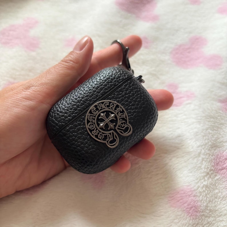 以下が含まれることがあります： シルバーのエンブレムが付いた黒革のAirPodケース。ケースはテクスチャ加工された表面で、キーホルダーやバッグに取り付けるための金属製の留め金が付いています。エンブレムは十字のデザインです。