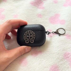 以下が含まれることがあります： 黒い革製AirPodケース。銀色の十字デザインのエンブレムが付いています。ケースは質感のある表面で、銀色の留め金が付いています。