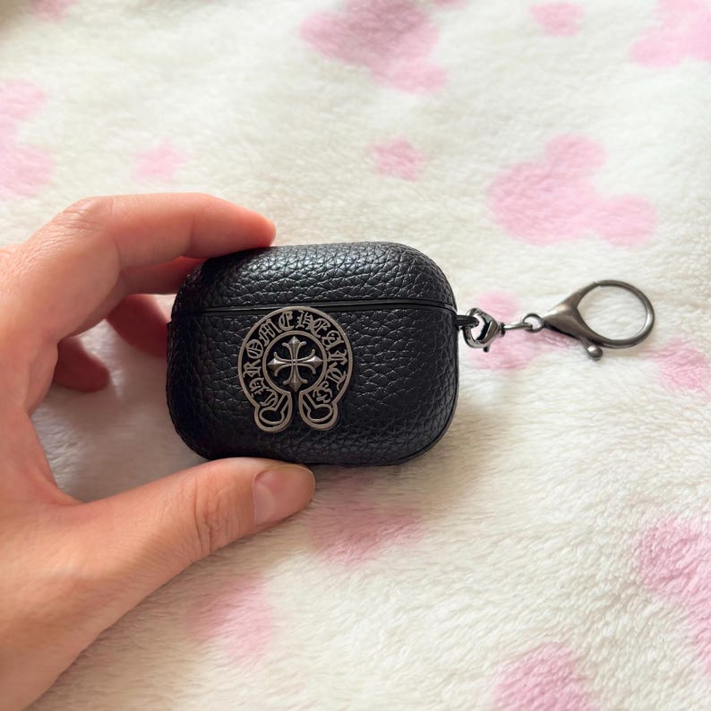 以下が含まれることがあります： 黒い革製AirPodケース。銀色の十字デザインのエンブレムが付いています。ケースは質感のある表面で、銀色の留め金が付いています。