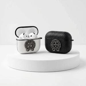 May include: Two AirPod cases, one white and one black, each with a decorative silver emblem. The white case is open, revealing white AirPods. The black case has a textured surface. Both cases are on a white platform.