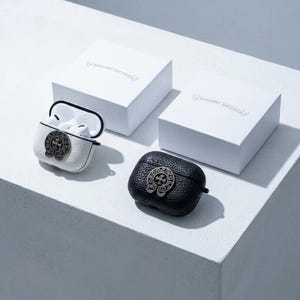 以下が含まれることがあります： シルバーのエンブレムが付いた、白と黒の革製AirPodsケース。 片方のケースは開いており、白いイヤホンが見えます。 エンボス加工されたテキストが入った白い箱が2つあり、パッケージを示唆しています。 ケースは白い表面にあります。