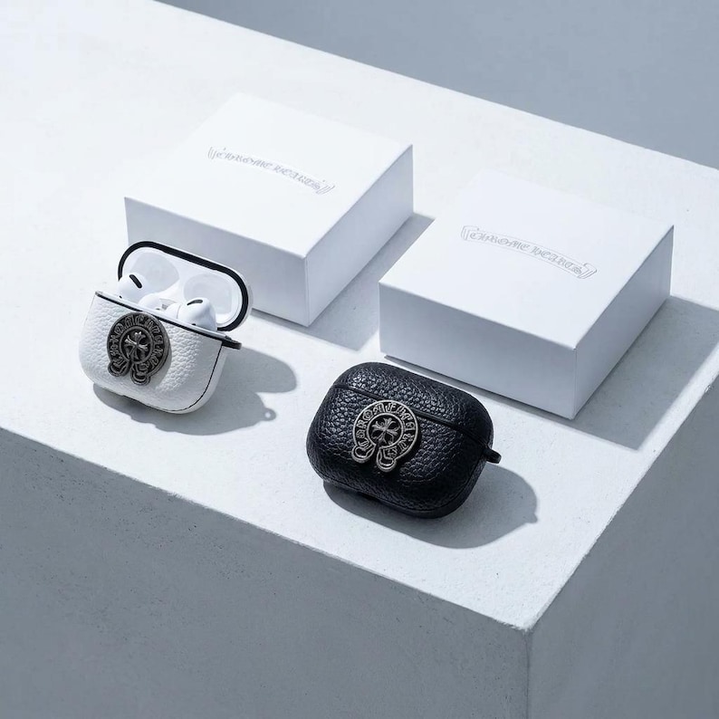 以下が含まれることがあります： シルバーのエンブレムが付いた、白と黒の革製AirPodsケース。 片方のケースは開いており、白いイヤホンが見えます。 エンボス加工されたテキストが入った白い箱が2つあり、パッケージを示唆しています。 ケースは白い表面にあります。