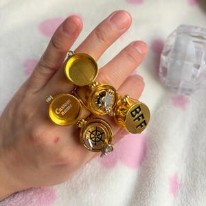 May include: Gold-colored rings with hinged compartments, some open to reveal small charms. One ring has "Custom name" text, another has "BFF" in black letters. The rings are displayed on a hand.