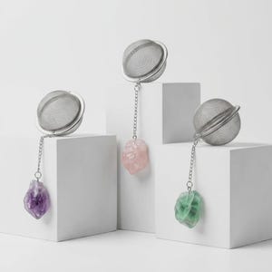 May include: Three stainless steel tea infusers with chain and gemstone accents. One features a purple amethyst, another a pink rose quartz, and the third a green fluorite. The infusers are displayed on white geometric blocks against a white background.