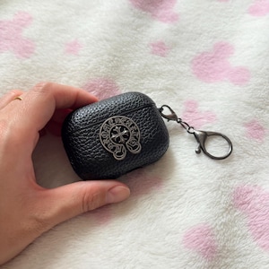 以下が含まれることがあります： 黒色の革製AirPodケース。銀色の十字デザインのエンブレムが付いています。ケースは質感のある表面で、持ち運びに便利なシルバー色のカラビナクリップが付いています。ワイヤレスイヤホンの保護と持ち運び用に設計されています。