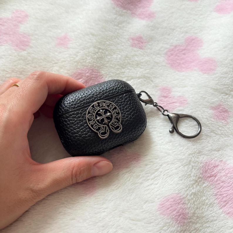 以下が含まれることがあります： 黒色の革製AirPodケース。銀色の十字デザインのエンブレムが付いています。ケースは質感のある表面で、持ち運びに便利なシルバー色のカラビナクリップが付いています。ワイヤレスイヤホンの保護と持ち運び用に設計されています。