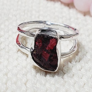 May include: A silver ring with a deep red, rough-cut gemstone. The ring has a double band design.