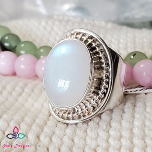 Puede incluir: Un anillo de plata con una piedra de luna blanca engastada en un engaste de alambre. El anillo est&aacute; sobre una superficie de punto blanco.