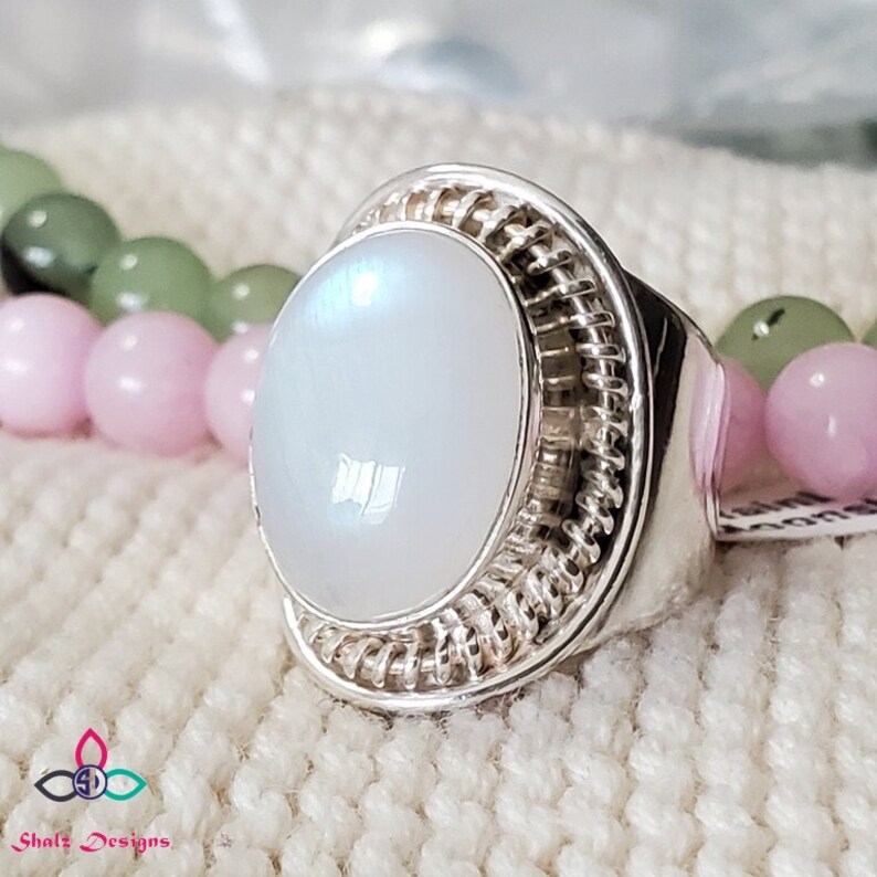Puede incluir: Un anillo de plata con una piedra de luna blanca engastada en un engaste de alambre. El anillo est&aacute; sobre una superficie de punto blanco.