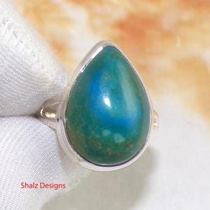 Op de afbeelding: Zilveren ring met een druppelvormige edelsteen met een blauw-groene kleurovergang. De ring heeft een gepolijste zilveren band en een bezel setting. De tekst "Shalz Designs" is zichtbaar. De steen is opvallend.