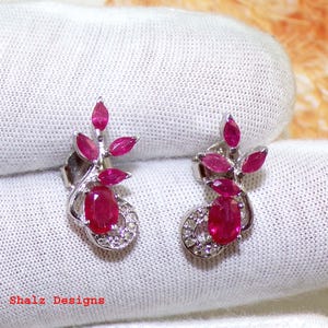 May include: A pair of silver earrings featuring red ruby gemstones. The earrings have a floral design with small ruby accents.