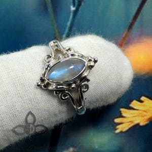 May include: A silver ring with a moonstone set in a detailed, ornate design. The ring is on a white fabric background.