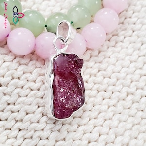 May include: A close-up of a silver pendant featuring a raw, pink ruby gemstone. The pendant is set against a background of pink and green beads.