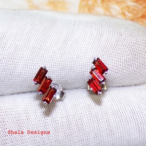 May include: A pair of silver earrings with red gemstone accents. The earrings feature a geometric design with three rectangular stones on each earring.