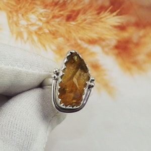 May include: A silver ring with a large, faceted, amber-colored gemstone set in a prong setting. The ring has a delicate, ornate design.