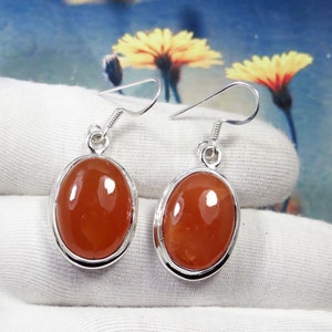 Könnte beinhalten: Ein Paar silberne Ohrringe mit orangefarbenen Karneol-Edelsteinen. Die Ohrringe haben eine einfache ovale Form und sind in einer silbernen Fassung gefasst.
