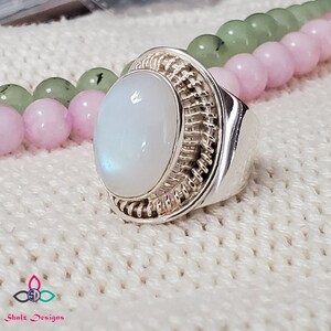 Puede incluir: Un anillo de plata con una gran piedra de luna ovalada engastada en una banda de plata tejida. El anillo est&aacute; sobre una superficie de punto blanco.