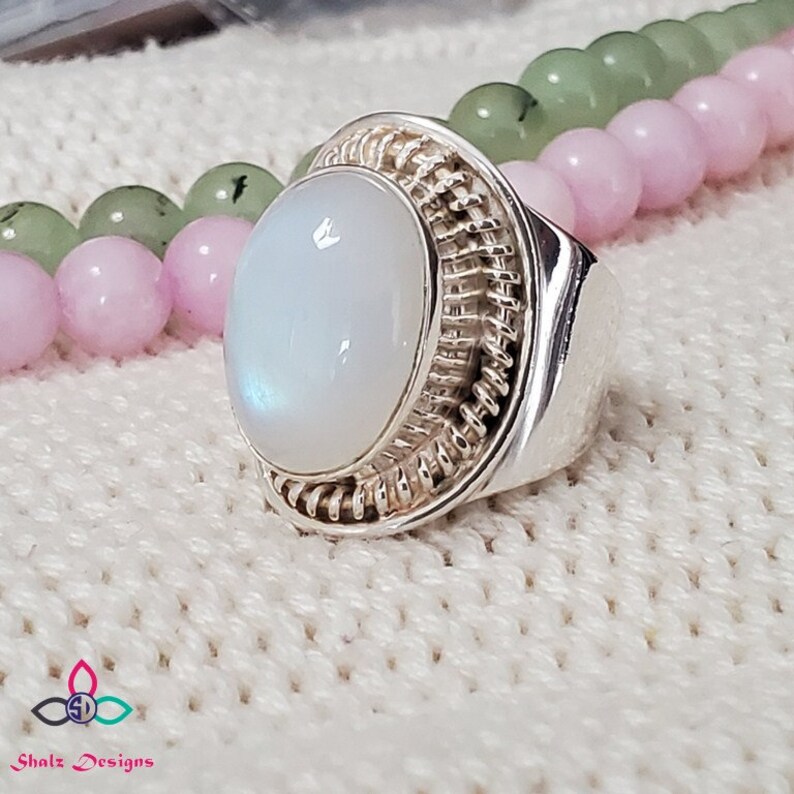 Puede incluir: Un anillo de plata con una gran piedra de luna ovalada engastada en una banda de plata tejida. El anillo est&aacute; sobre una superficie de punto blanco.