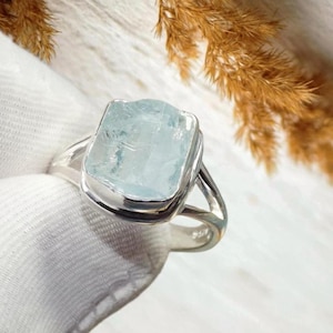 May include: A silver ring featuring a large, rectangular aquamarine gemstone. The pale blue-green stone is set in a bezel setting with a split shank design. The ring is displayed on a white surface.