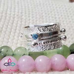 May include: A silver spinning ring with a blue gemstone set in the centre. The ring has a textured design and is surrounded by a row of beads.