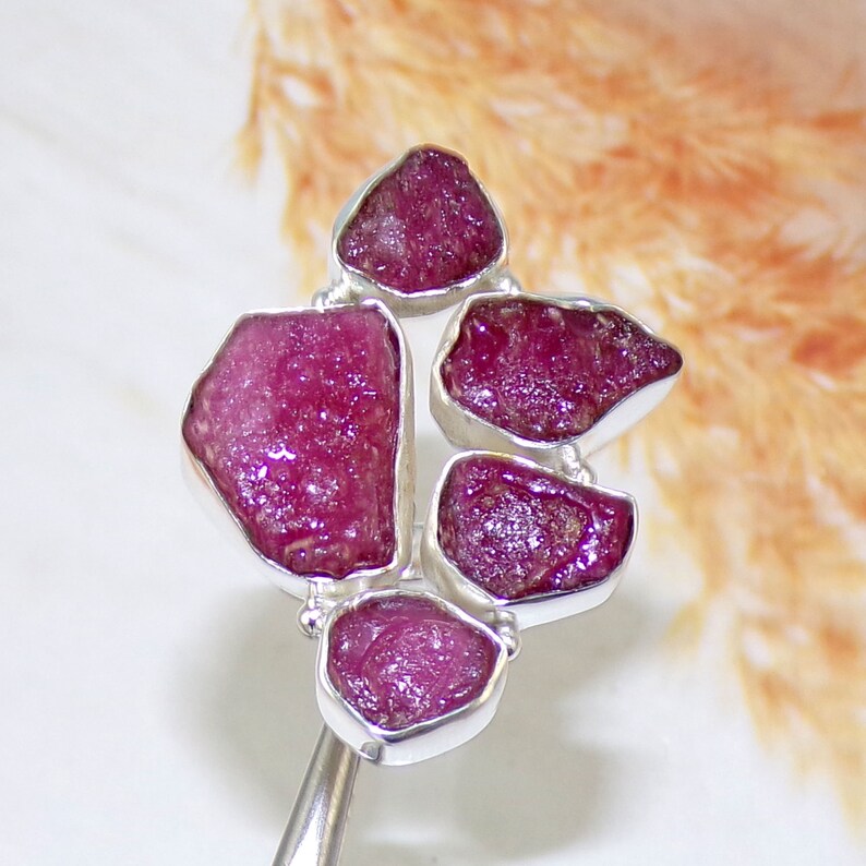 May include: A silver ring with six raw ruby gemstones set in a cluster. The rubies are a deep pink color and have a rough, natural texture.