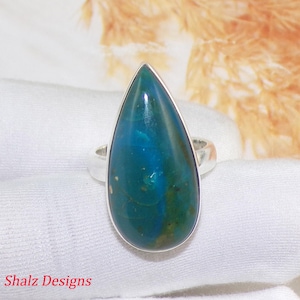 Op de afbeelding: Een zilveren ring met een druppelvormige, gepolijste steen in blauwgroene tinten. De steen heeft een wervelend patroon. De ring ligt op een witte stof. De tekst "Shalz Designs" is zichtbaar.