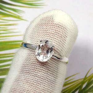 May include: A silver ring featuring a clear oval-shaped gemstone. The ring is displayed on a white fabric background.