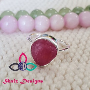 May include: A silver ring with a pink gemstone set in a bezel setting. The ring is on a white textured surface.