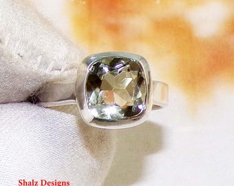 Puede incluir: Un anillo de plata con una gema grande, de corte cuadrado y verde pálido. La gema está engastada en un bisel, y la banda del anillo es sencilla y pulida. El anillo se muestra sobre una superficie blanca, con el texto "Shalz Designs".