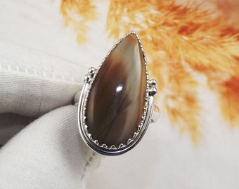 Könnte beinhalten: Ein tropfenförmiger Ring mit einem braunen und weißen Achat-Edelstein, der in einer silbernen Fassung sitzt. Der Ring hat einen zarten, gewellten Rand.