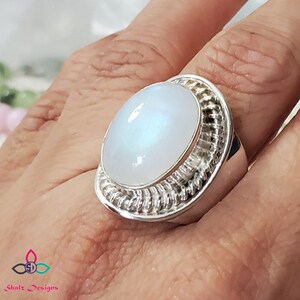 Puede incluir: Un anillo de plata con una gran piedra de luna ovalada engastada en un engaste de alambre. El anillo est&aacute; en un dedo.