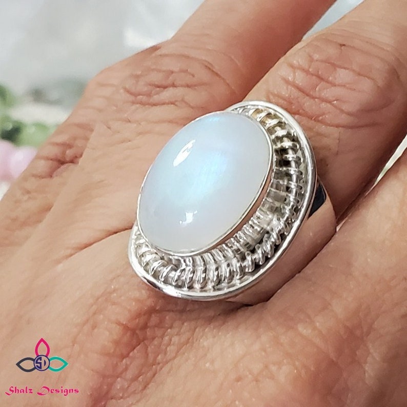 Puede incluir: Un anillo de plata con una gran piedra de luna ovalada engastada en un engaste de alambre. El anillo est&aacute; en un dedo.