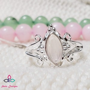 May include: A silver ring with a delicate, ornate design features a marquise-shaped, milky white gemstone. The ring is set against a background of pink and green beads.