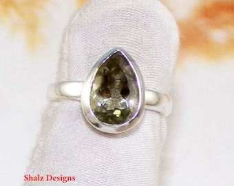 Puede incluir: Un anillo de plata con una piedra preciosa verde oliva en forma de pera. La piedra está engastada en un bisel y la banda es lisa y pulida. El anillo se muestra sobre una tela blanca.