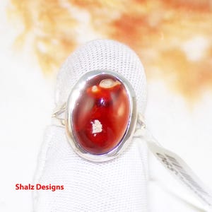 Può includere: Un anello d'argento con una pietra ovale rossa e bianca. L'anello è su uno sfondo di tessuto bianco.