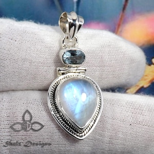 May include: A silver pendant with a teardrop-shaped moonstone and a smaller oval-shaped blue topaz. The pendant has a detailed silver border.