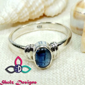 May include: A silver ring featuring a blue oval gemstone. The ring has a plain band with decorative detailing either side of the gemstone. The ring is set against a soft, neutral background.