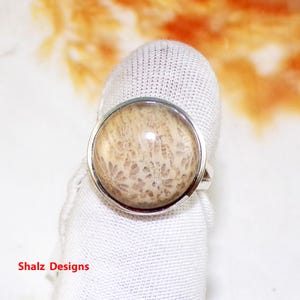 Pode incluir: Um anel de prata com uma pedra redonda de coral fossilizado de cor castanho claro engastada no centro. A pedra tem um padrão detalhado e intrincado.