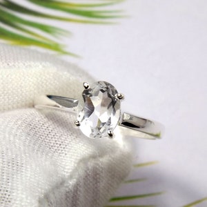 May include: A silver ring featuring a clear oval-shaped gemstone. The ring is displayed on a white fabric background.