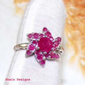 May include: A silver ring with a pink ruby flower design. The flower has a large oval ruby in the center and smaller rubies surrounding it.
