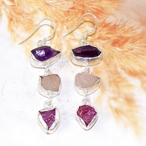 May include: Dangle earrings featuring three raw gemstone pieces in purple, pink, and red hues. The stones are set in silver-colored metal. The earrings have a hook closure.