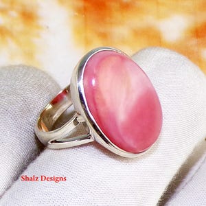 May include: A silver ring with a large, oval pink gemstone. The gemstone has a polished surface with a gradient of pink shades. The ring's band splits at the top, creating a distinctive design. The ring is set on a white surface.