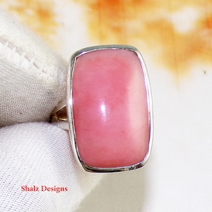 May include: A silver ring with a large, rectangular pink gemstone. The gemstone has a smooth, polished surface and is set in a silver bezel. The ring is displayed on a white surface. The text "Shalz Designs" is visible.