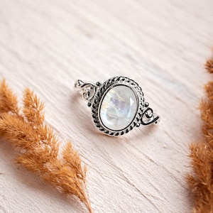 May include: A silver ring featuring an oval-shaped, iridescent moonstone cabochon set within a twisted rope-style metal border. The band displays decorative scrollwork details on the sides, framing the central gemstone with a polished finish.
